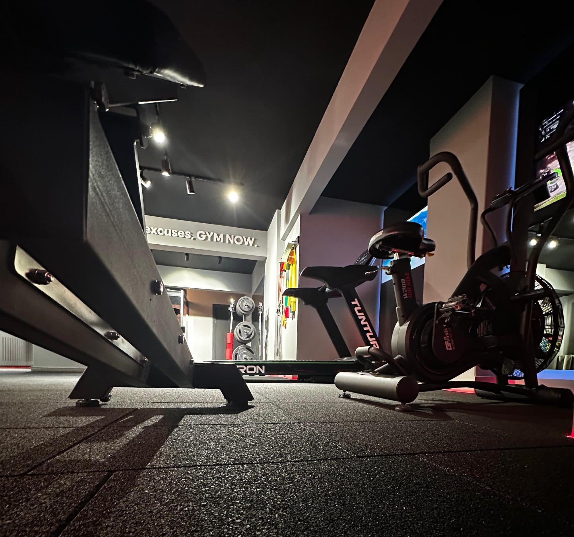 Yer seviyesinden çekilmiş kardiyo alanı; air bike, koşu bandı ve arkada ağırlık plakaları